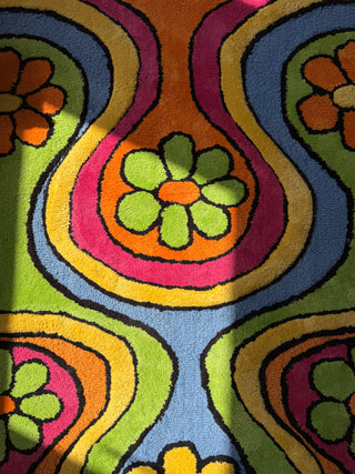 A close-up of a colorful retro-style tufted rug or textile with bold wavy bands and flower motifs in green, orange, pink, yellow, blue, and black.